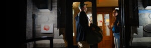 Man carrying a large duffel bag talks to a smiling woman inside a doorway at a brick building entrance at night.