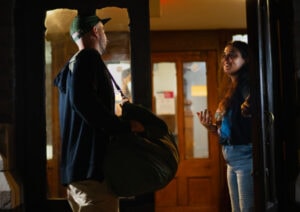 Man carrying a large duffel bag speaks with a woman at a warmly lit doorway in a hallway.